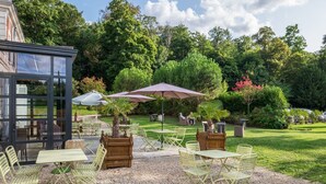 Een buitenterras met tafels en stoelen, omgeven door groen en bomen.