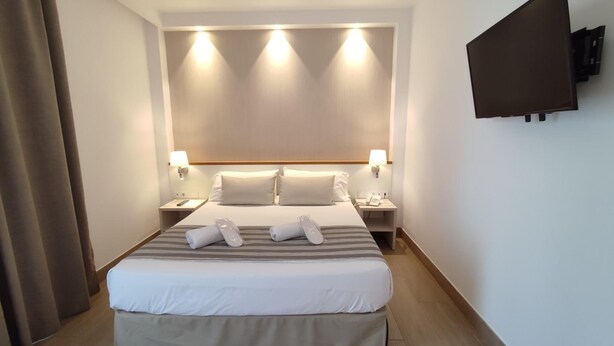 Habitación de hotel con una cama grande, dos mesitas de noche, un televisor de pantalla plana montado en la pared y una cortina a la izquierda.