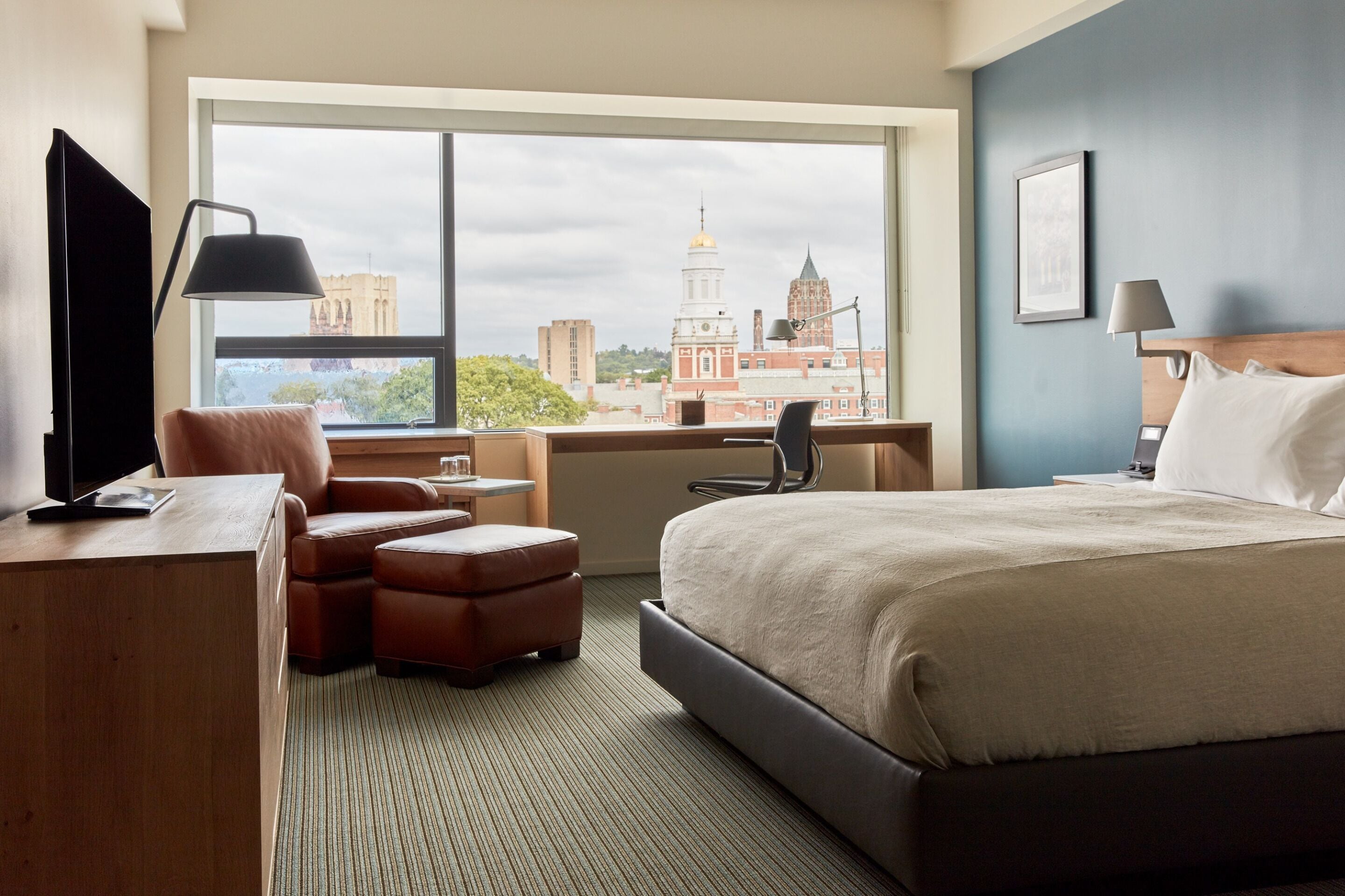 A hotel room with a large window, a bed, a desk, a chair, and a TV.