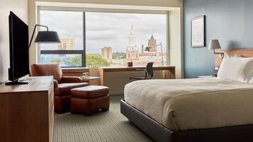 A hotel room with a large window, a bed, a desk, a chair, and a TV.