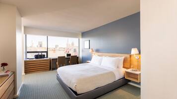 Quarto de hotel com uma cama grande, uma escrivaninha, uma cadeira, vista para a cidade e uma mesa de cabeceira com abajur.