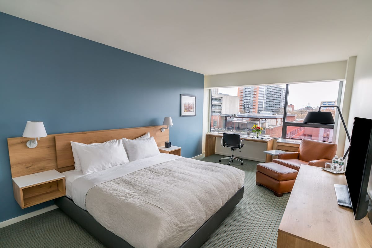 A hotel room with a large bed, a desk, a chair, a TV, and a window with a city view.