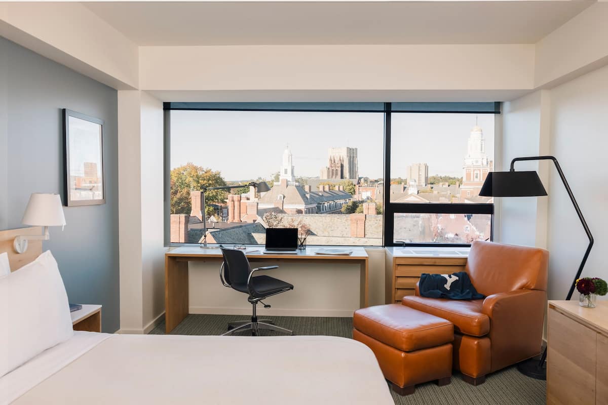 A hotel room with a bed, desk, chair, and a large window with a city view.