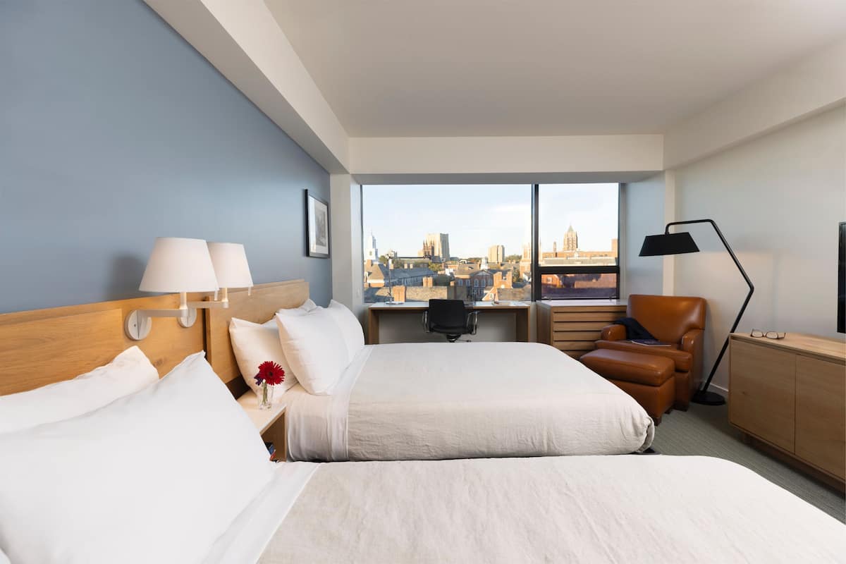 A hotel room with two beds, a desk, a chair, a lamp, and a view of a cityscape.