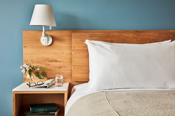 A neatly made bed with white bedding, a wooden headboard, and a bedside table with a lamp, books, and a vase with flowers.