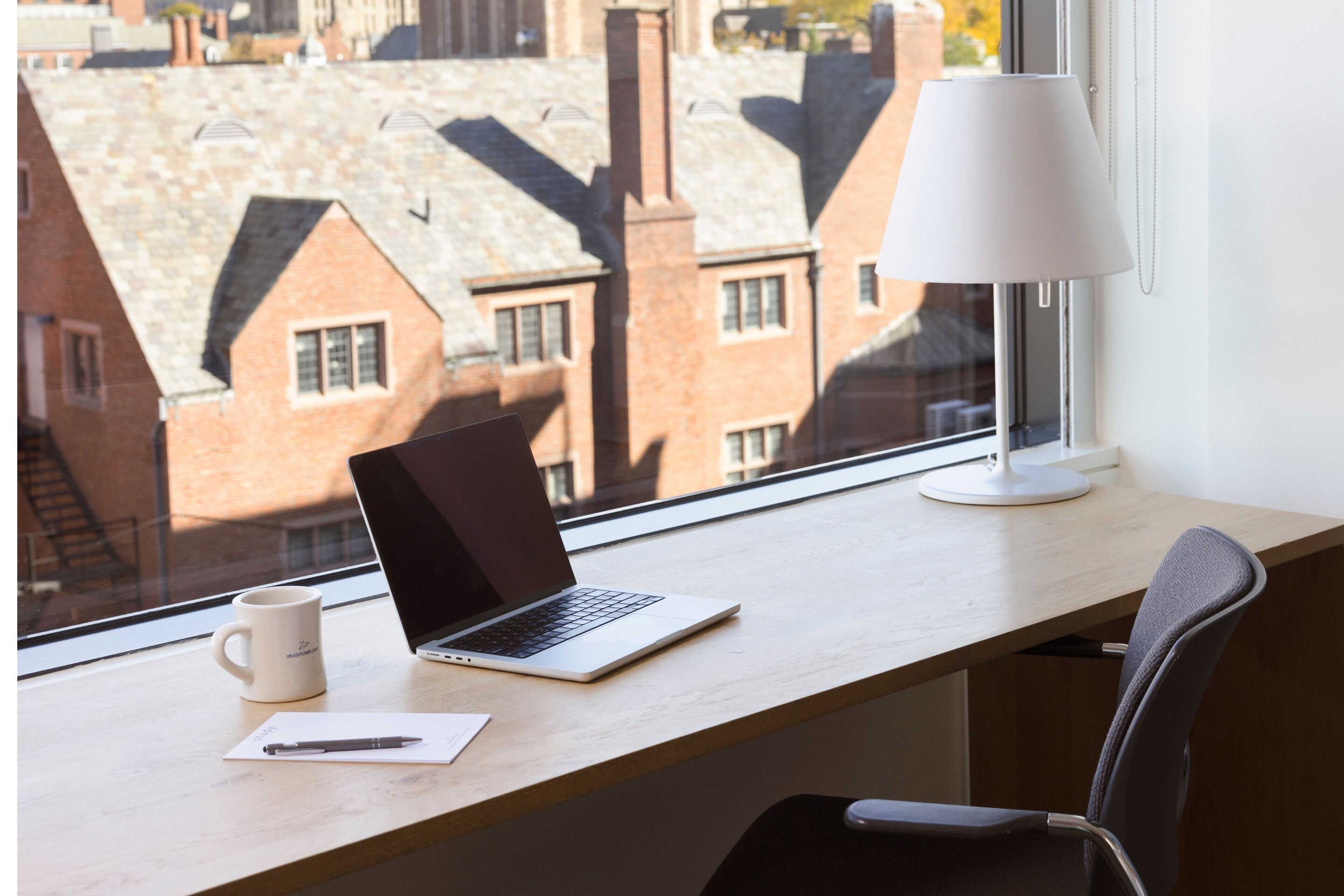 Una laptop sobre un escritorio junto a una ventana, con una lámpara, una taza y una silla.