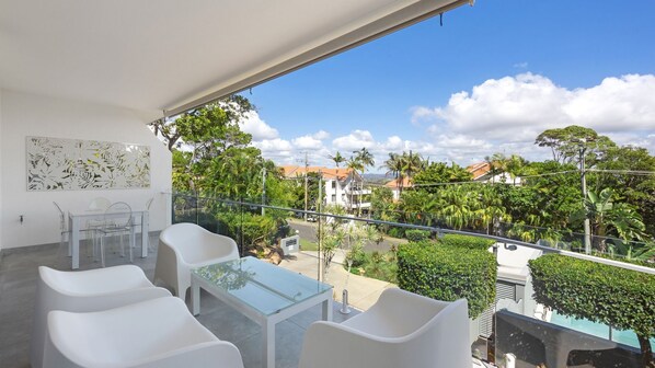 Un balcón con una mesa de vidrio y sillas blancas, con vistas a una zona residencial con palmeras y casas.