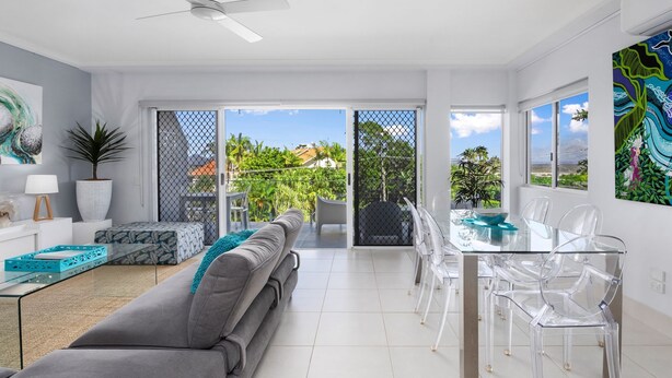 A modern living room with a grey sofa, a glass dining table with clear chairs, a ceiling fan, and a view of a balcony with plants and a fence.