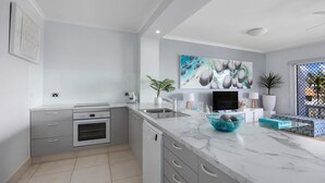 A modern kitchen with grey cabinetry, a built-in oven, a dishwasher, and a marble countertop. There is a large painting above the TV, and a potted plant on the counter.