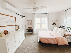 Une chambre à coucher comprenant un lit, une commode, un ventilateur de plafond, une fenêtre avec des rideaux et un mur orné d’une citation inspirante.