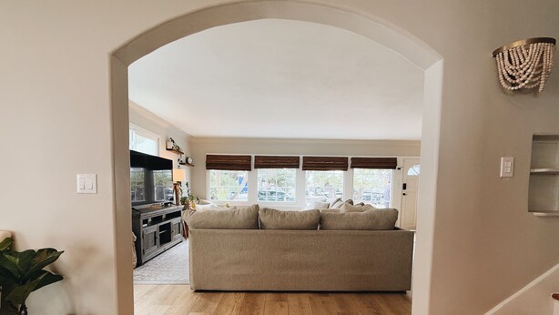 A living room with a beige sofa, wooden flooring, and a large window with blinds.