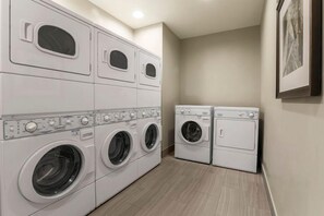 Une buanderie équipée de machines à laver et de sèche-linge superposés, avec un sol carrelé et un tableau encadré accroché au mur.