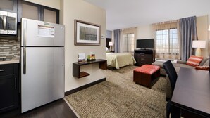 Ein kompaktes Hotelzimmer mit Küchenzeile, einem Bett mit grüner Bettdecke, einem Fernseher und einer Sitzecke mit Tisch und Stühlen.