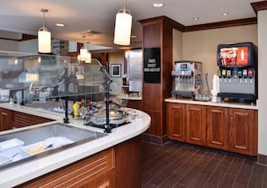 A breakfast bar with a glass partition, a Coca-Cola dispenser, and a sign offering free daily breakfast.
