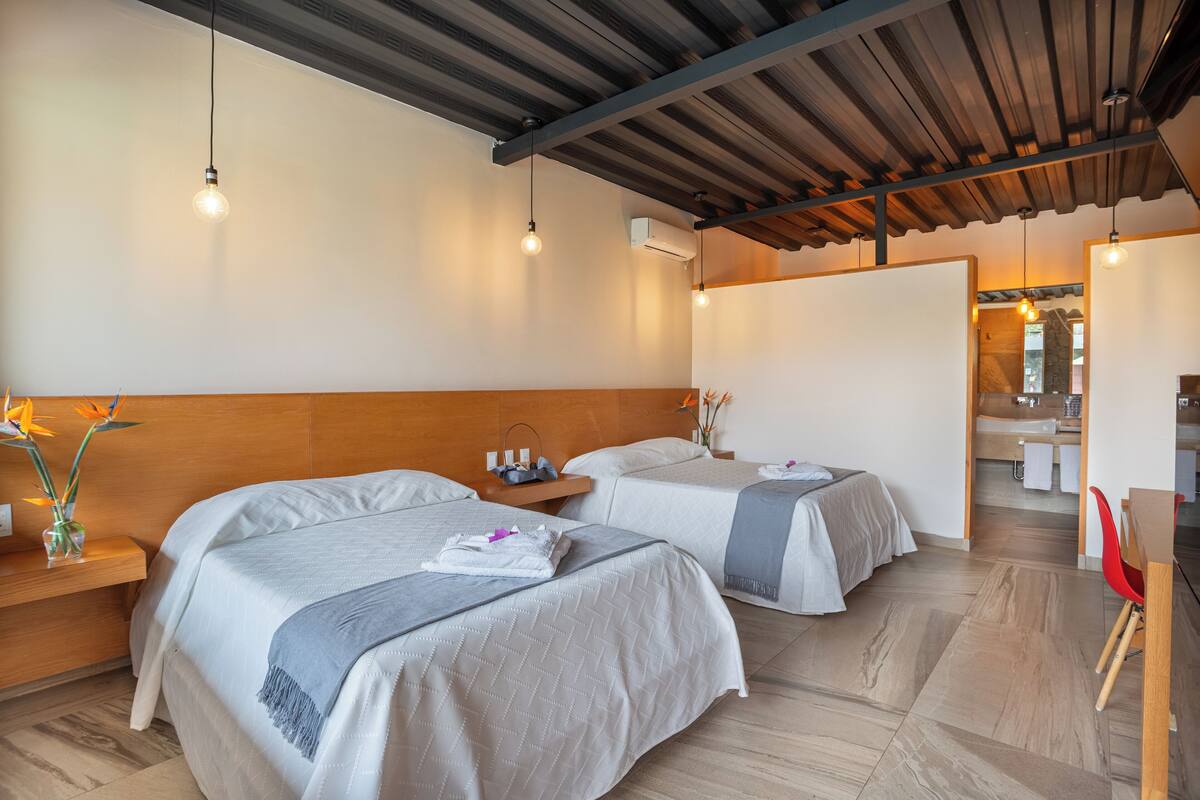 Une chambre d’hôtel moderne avec deux lits, des têtes de lit en bois, un plafond en bois et une salle de bain visible à travers une porte ouverte.