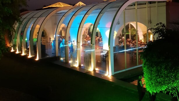 Una estructura moderna con paredes de vidrio y varios niveles, posiblemente un restaurante o espacio para eventos, con gente dentro y fuera.