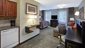 A hotel room with a kitchenette, a bed, a desk, a chair, a TV, and a window with curtains.