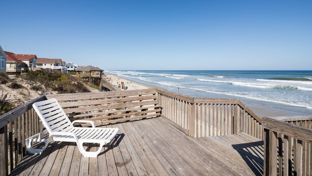 Een houten terras met twee witte ligstoelen met uitzicht op een strand met golven en huizen op de achtergrond.