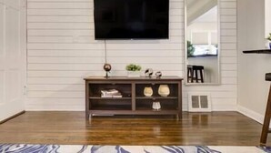 A living room with a wooden floor, a TV mounted on the wall, a dark wooden console table with decorative items, and a large mirror reflecting part of the room. - Walk to Falls / Close to Everything / Fenced Backyard (Minneapolis)