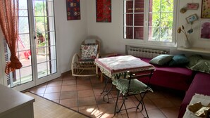 A room with a large sliding glass door, a wicker chair with a floral cushion, a small table with a tablecloth, and a purple sofa with cushions. - At 8km from the sea two-story apartment in villa (Cisano Sul Neva)