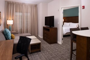Una camera d'hotel con un letto, una TV su un supporto in legno, una scrivania con una sedia, un divano con dei cuscini e un tavolino.