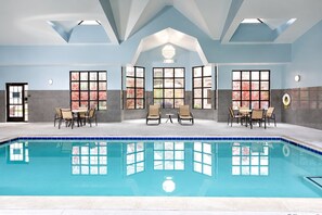 Une piscine intérieure avec un sol carrelé, entourée d’espaces aménagés pour s’asseoir et dotée de grandes fenêtres.