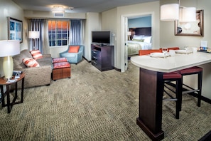 Une chambre d’hôtel équipée d’un canapé, d’un fauteuil, d’un pouf, d’une télévision, d’un bureau et d’un coin repas.