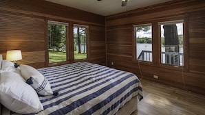 A bedroom with a bed, a ceiling fan, and large windows offering a view of a lake and trees.