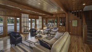 A spacious living room with a wooden ceiling, a sofa set, a coffee table, and a view of a lake through large glass doors.
