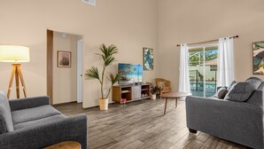 A spacious living room with a grey sofa, a wooden coffee table, and a TV stand. There are potted plants and artwork on the walls. - Amazing Blue Private Pool - 4BR House Near Disney (Kissimmee)