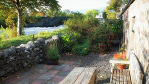 Eine Steinsäule, eine Holzbank und Kübelpflanzen in einem Garten mit Blick auf einen Fluss.