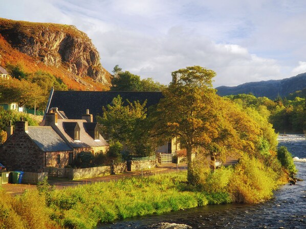 A stone house with a sloped roof near a river, surrounded by lush greenery and a rocky cliff.