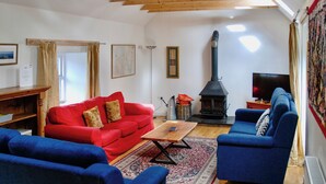 Ein Wohnzimmer mit einem roten Sofa, blauen Sesseln, einer Holzbalkendecke, einem schwarzen Holzofen und einem Fernseher.