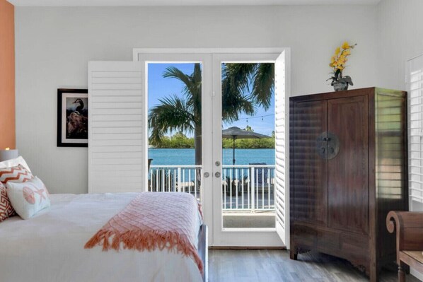 A bedroom with a bed, a wooden cabinet, and a door leading to a balcony with a view of the sea and palm trees.