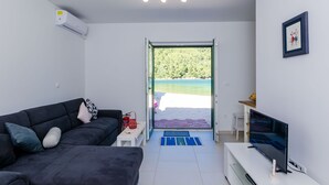 Una sala de estar moderna con un sofá oscuro, un televisor de pantalla plana y vista a un paisaje verde a través de un ventanal.
