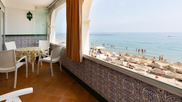Un balcón con vista a la playa, sillas de mimbre blancas, una mesa con mantel de colores y azulejos decorativos en las paredes.