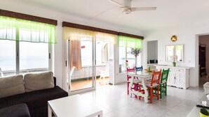 Un salón luminoso con un sofá, una mesa de comedor con sillas de colores, y una zona de cocina con un armario blanco.