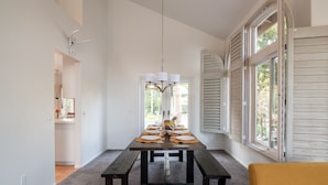 A dining area with a wooden table set for six, a chandelier, and a view of the outdoors through large windows. - Sunset Disco: Large Pool/Spa @ Lush Family Retreat (La Quinta)