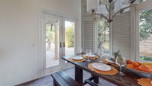 A dining area with a wooden table set for four, a bench, and a view of the garden through the door. - Sunset Disco: Large Pool/Spa @ Lush Family Retreat (La Quinta)