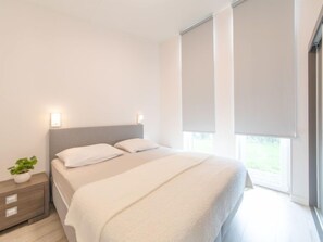 A modern bedroom with a large bed, a bedside table with a plant, and a window with blinds.