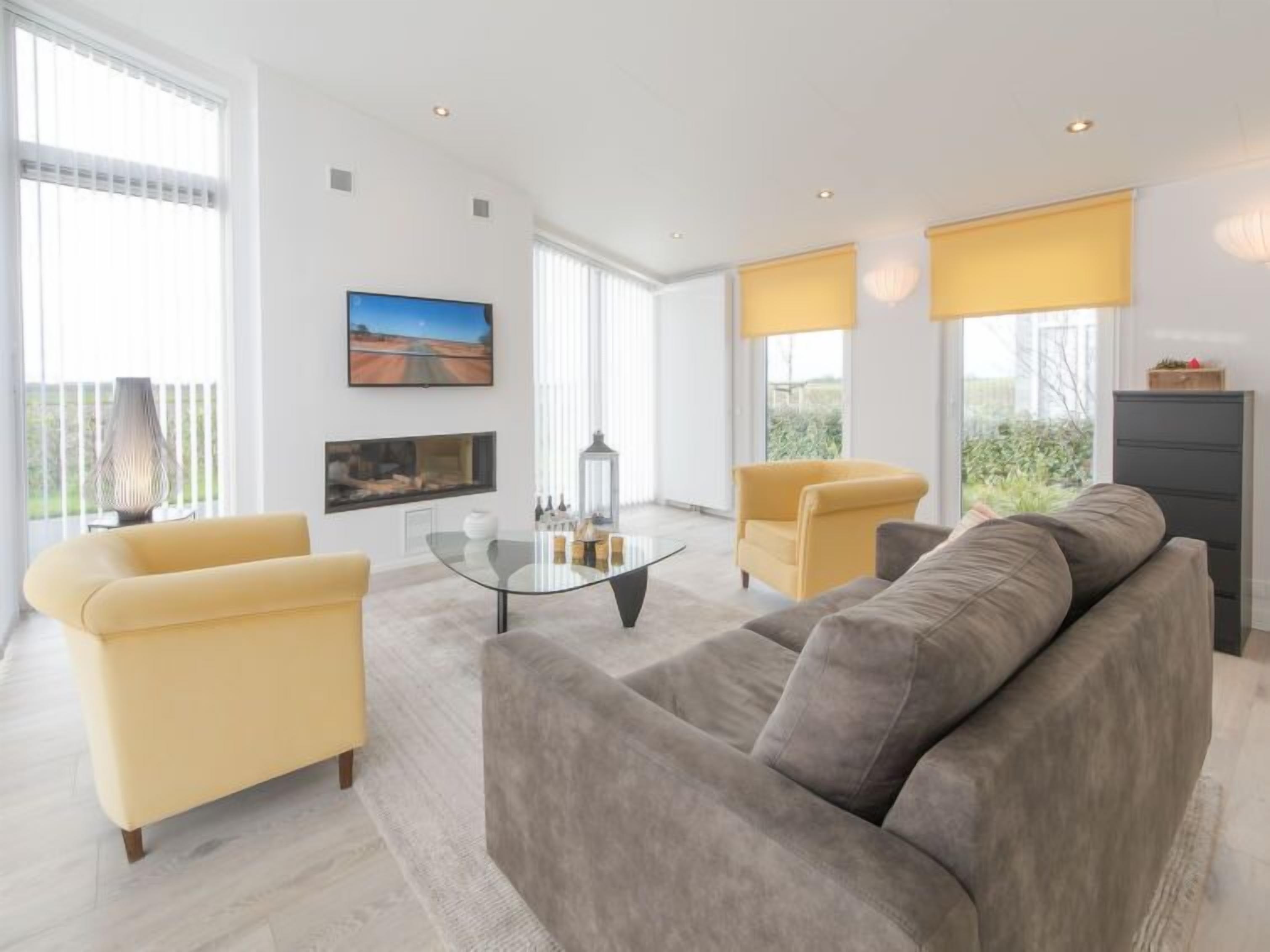 Un salon moderne avec un canapé gris, des fauteuils jaunes, une table basse en verre et un téléviseur à écran plat fixé au mur.