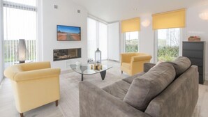 Una sala de estar moderna con un sofá gris, sillones amarillos, una mesa de centro de vidrio y un televisor de pantalla plana montado en la pared.