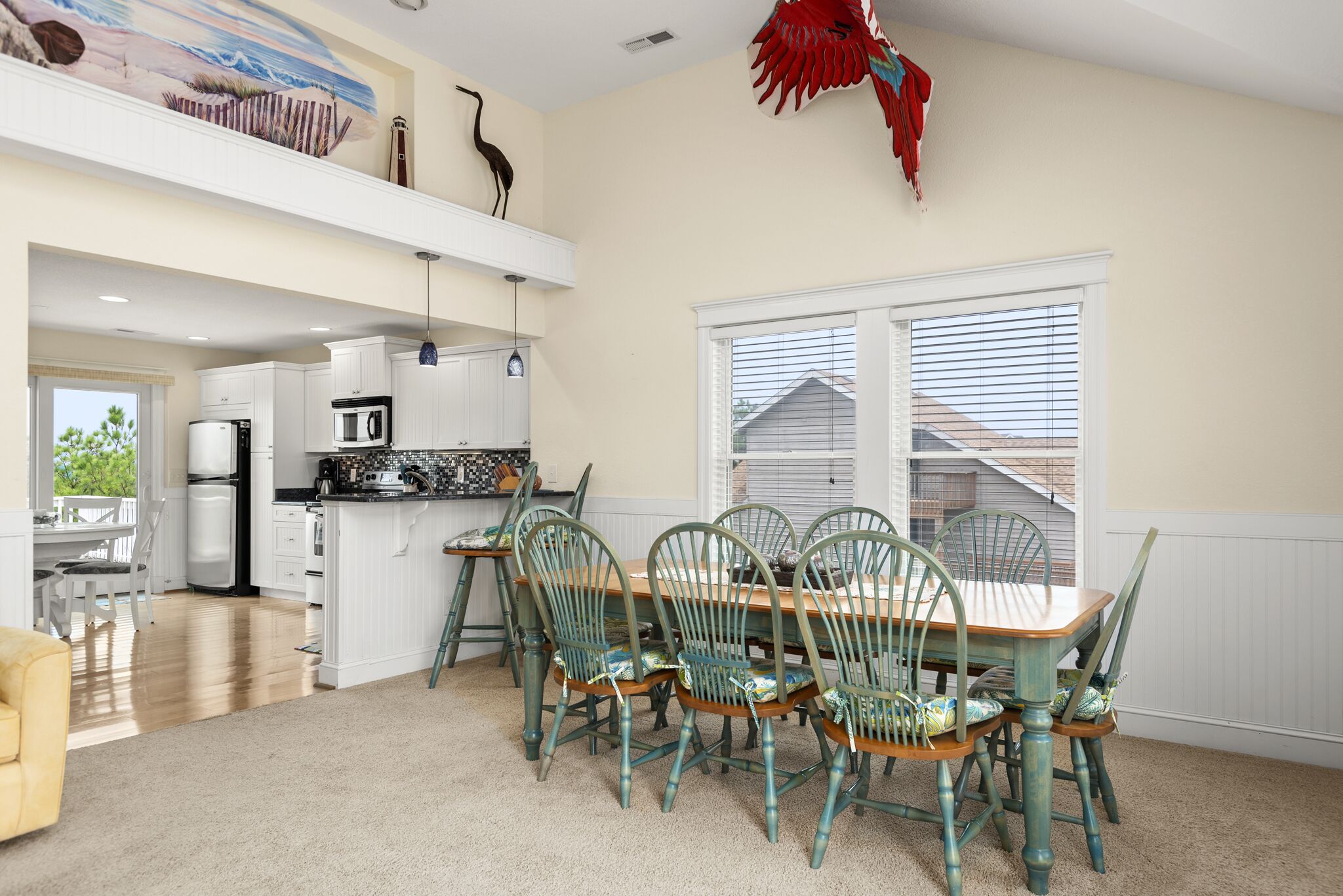 A dining area with a wooden table and green chairs, a kitchen with white cabinets, and a large window with blinds.