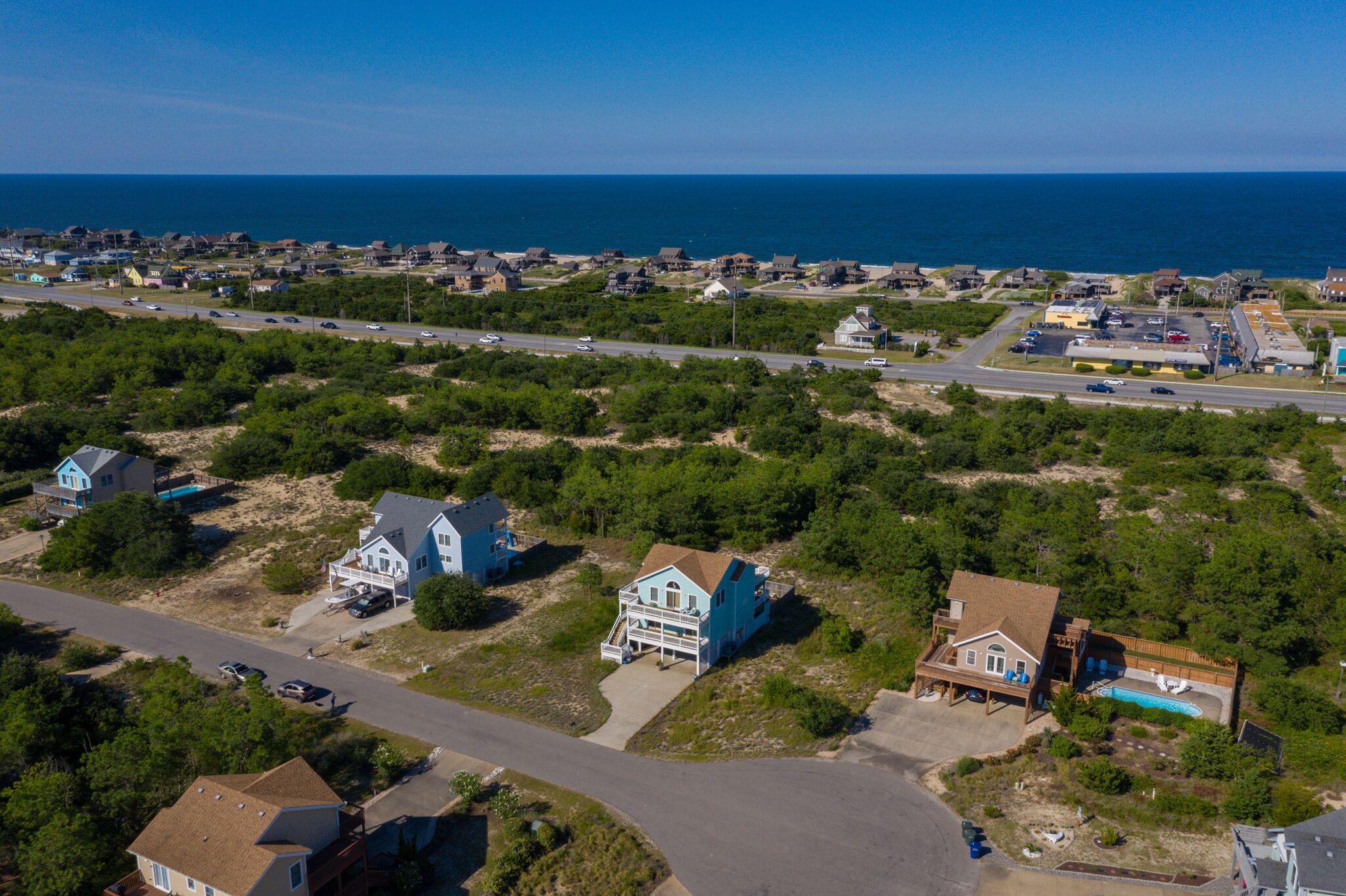 An aerial view of a coastal residential area with houses, a road, and a beach in the background.