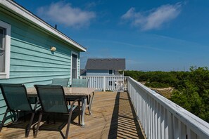 Um deck de madeira com uma mesa e cadeiras, um corrimão branco e vista para uma casa com fachada azul e telhado cinzento.