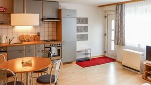 Een kleine keuken met moderne apparatuur, een eethoek met een tafel en stoelen, en een woonruimte met een televisie en een raam met gordijnen.