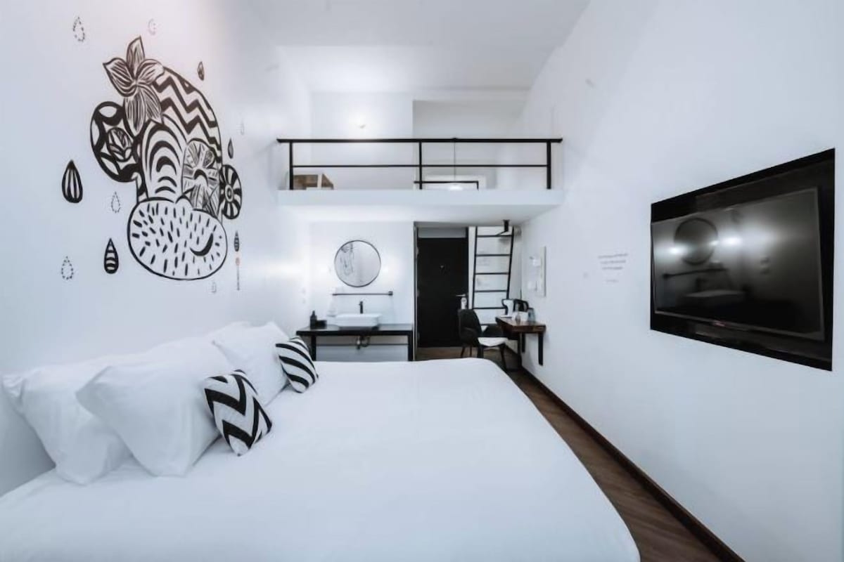 A modern hotel room with a large bed, a TV mounted on the wall, a desk with a chair, and a small kitchenette.