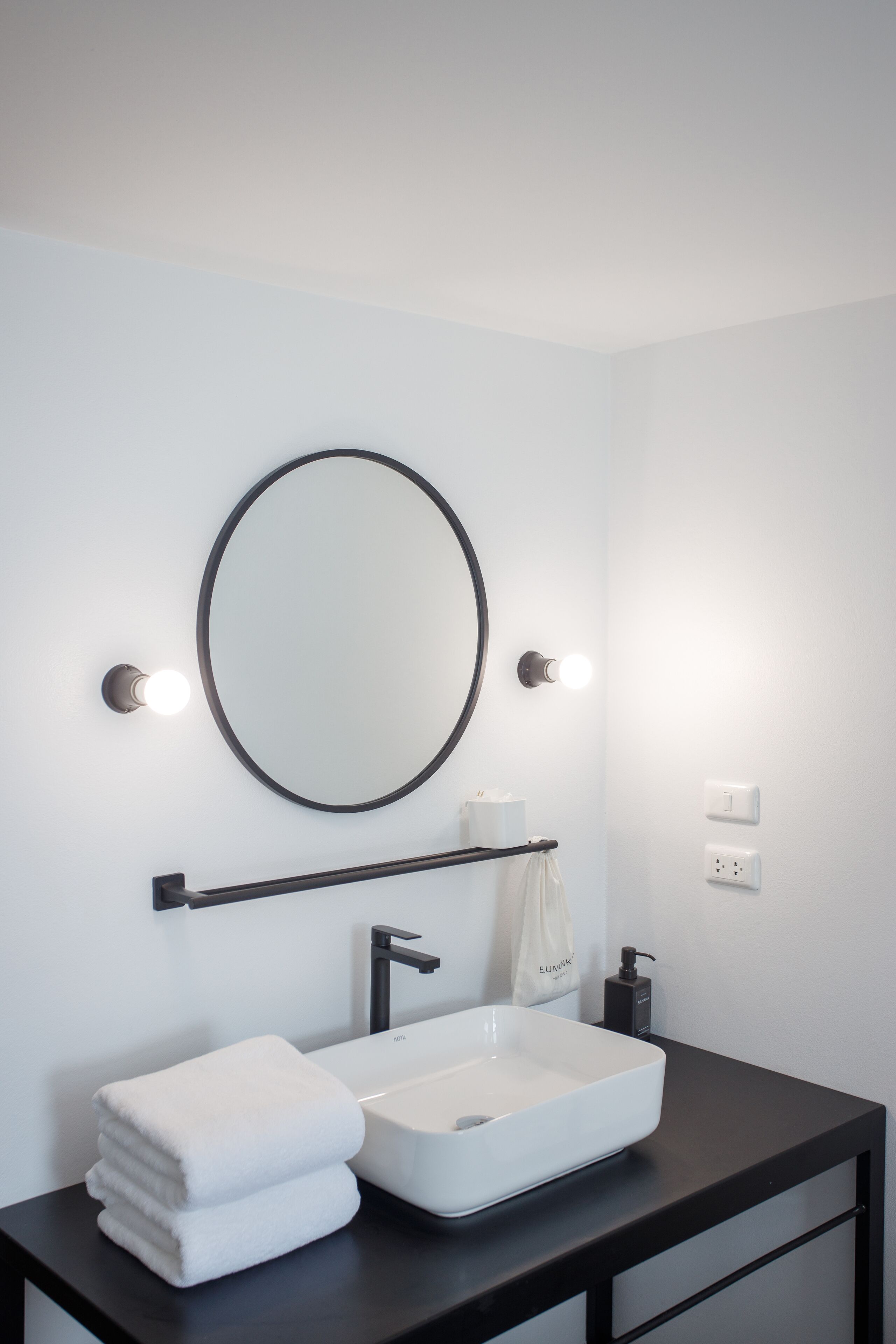 A modern bathroom with a black countertop, a white sink, a round mirror, and two wall-mounted lights.