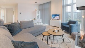 Ein modernes Wohnzimmer mit einem großen grauen Sofa, einem kleinen runden Tisch und einem blauen Sessel. An der Wand ist ein Fernseher angebracht und es gibt einen Kamin mit brennendem Holz.