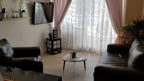 A living room with a sofa, a coffee table, and a TV mounted on the wall. There are potted plants on a shelf and a speaker on a stand. - Apartment in front of La Herradura Beach ideal for the Family (Coquimbo)